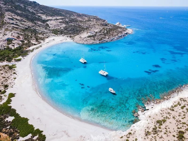 An aerial view of boats in a small bay on the island Milos in Greece, surrounded by turquoise waters all around.